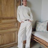 Person wearing a white sweater and pants standing in a room with wooden door and floral-patterned cushion.