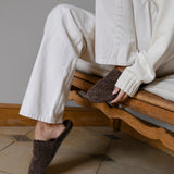 Person wearing brown mules sitting on a wooden chair indoors.