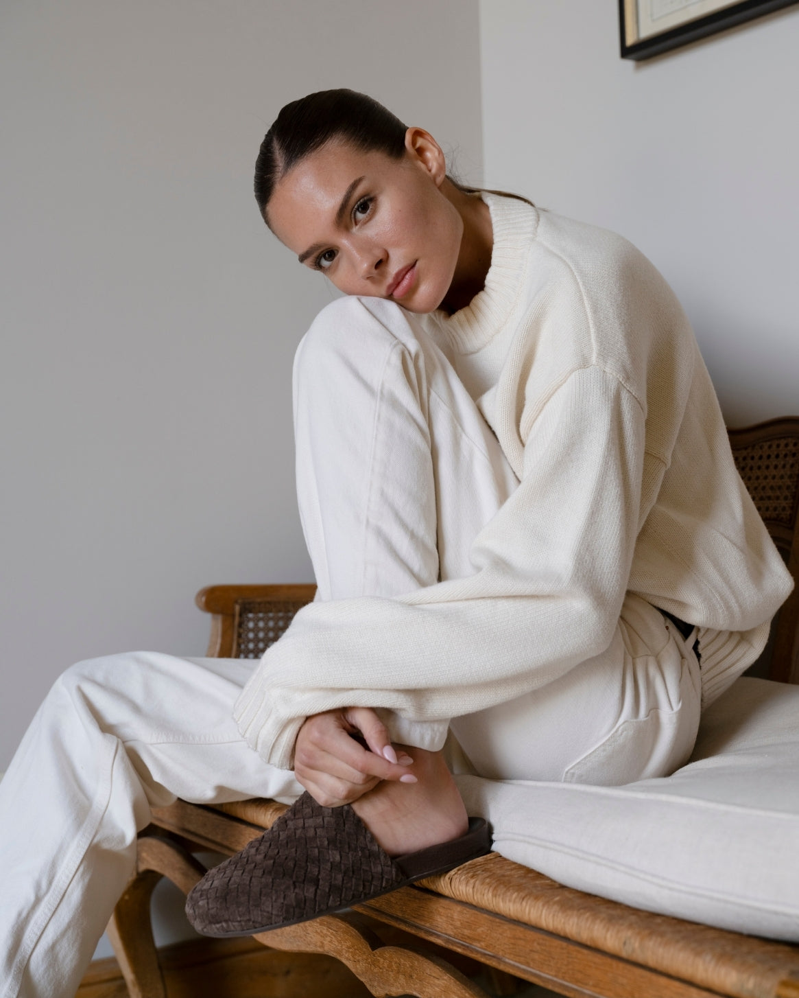 A woman sits on a wooden chair wearing an oversized cream knit sweater and matching cream sweatpants, with one brown textured mule visible.