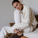 A woman sits on a wooden chair wearing an oversized cream knit sweater and matching cream sweatpants, with one brown textured mule visible.