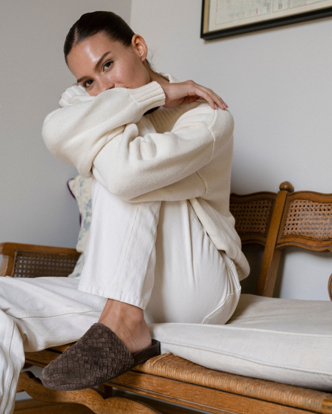 Woman wearing a white sweater and white pants sitting on a wooden bench indoors.