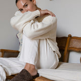 Woman wearing a white sweater and white pants sitting on a wooden bench indoors.