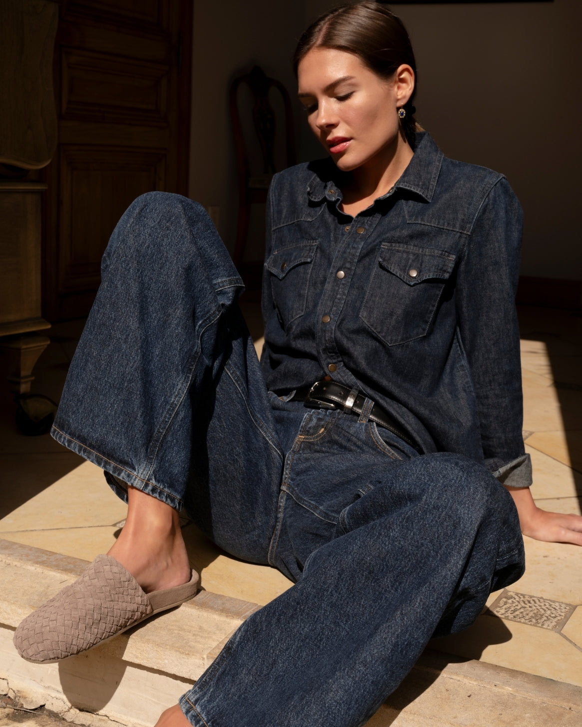 A woman sits on stone steps wearing a dark blue denim button-up shirt and matching wide-leg jeans, with a black belt and woven taupe slip-on shoes.