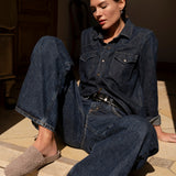 A woman sits on stone steps wearing a dark blue denim button-up shirt and matching wide-leg jeans, with a black belt and woven taupe slip-on shoes.