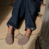 A person sitting on a tiled floor wearing dark blue wide-leg jeans and taupe woven slip-on shoes.