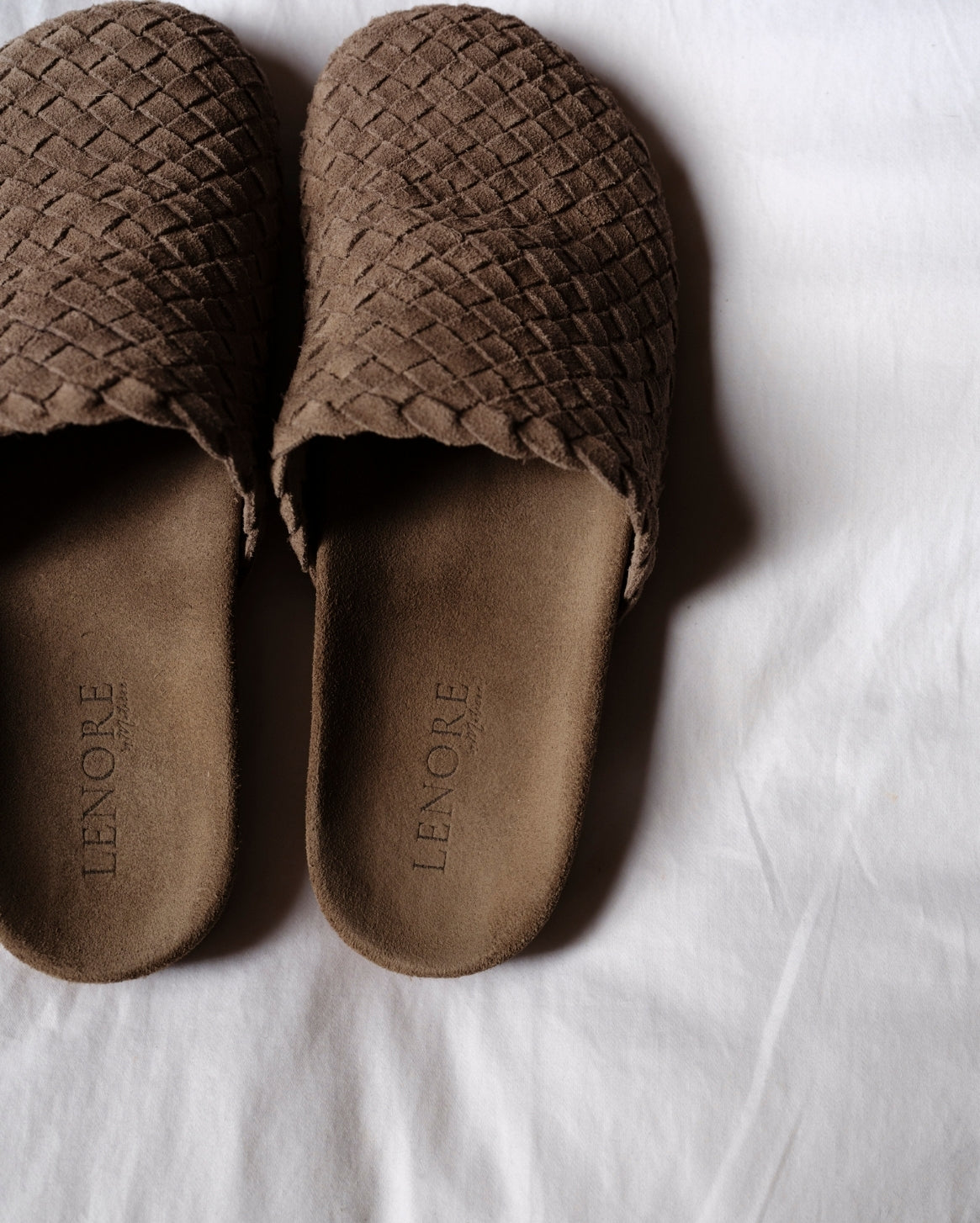 A pair of brown woven slip-on shoes with a suede-like footbed, laid flat on a white fabric background. The brand name 'LENORE' is debossed on the footbed.