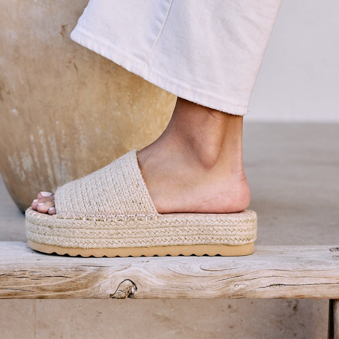 Ivory platform slide sandal with woven jute upper and thick sole, shown on a wooden bench