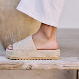 Ivory platform slide sandal with woven jute upper and thick sole, shown on a wooden bench