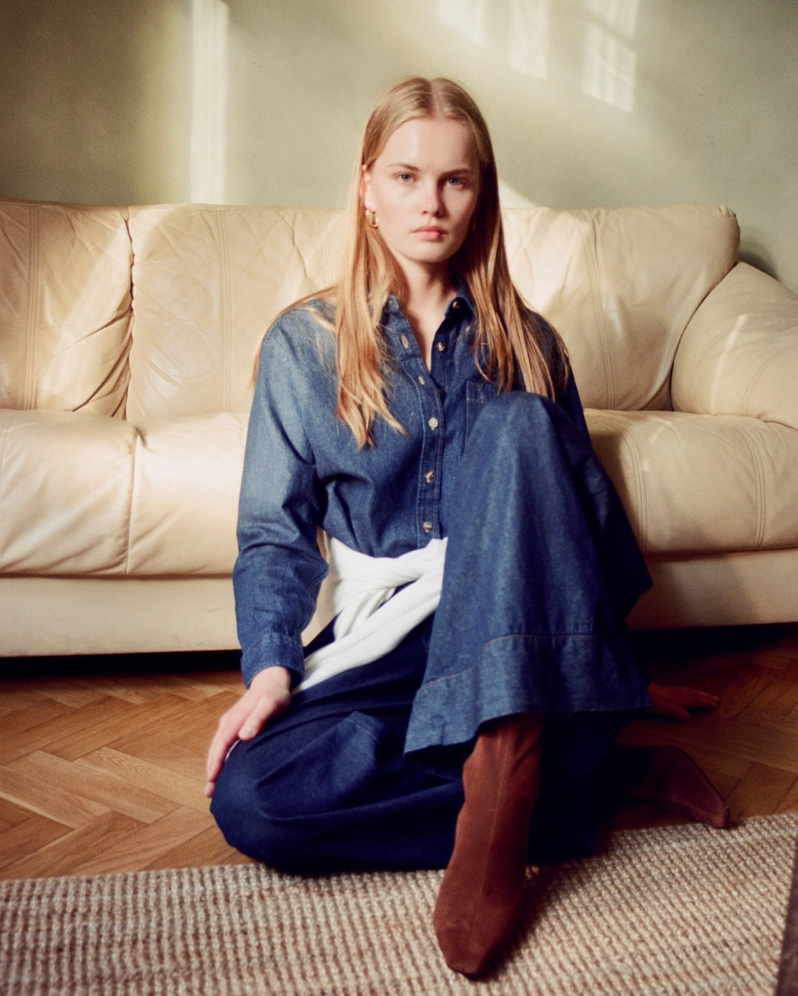 A woman sits cross-legged on a woven rug in front of a cream leather sofa, wearing a loose blue denim shirt and matching wide-leg denim pants with brown boots.