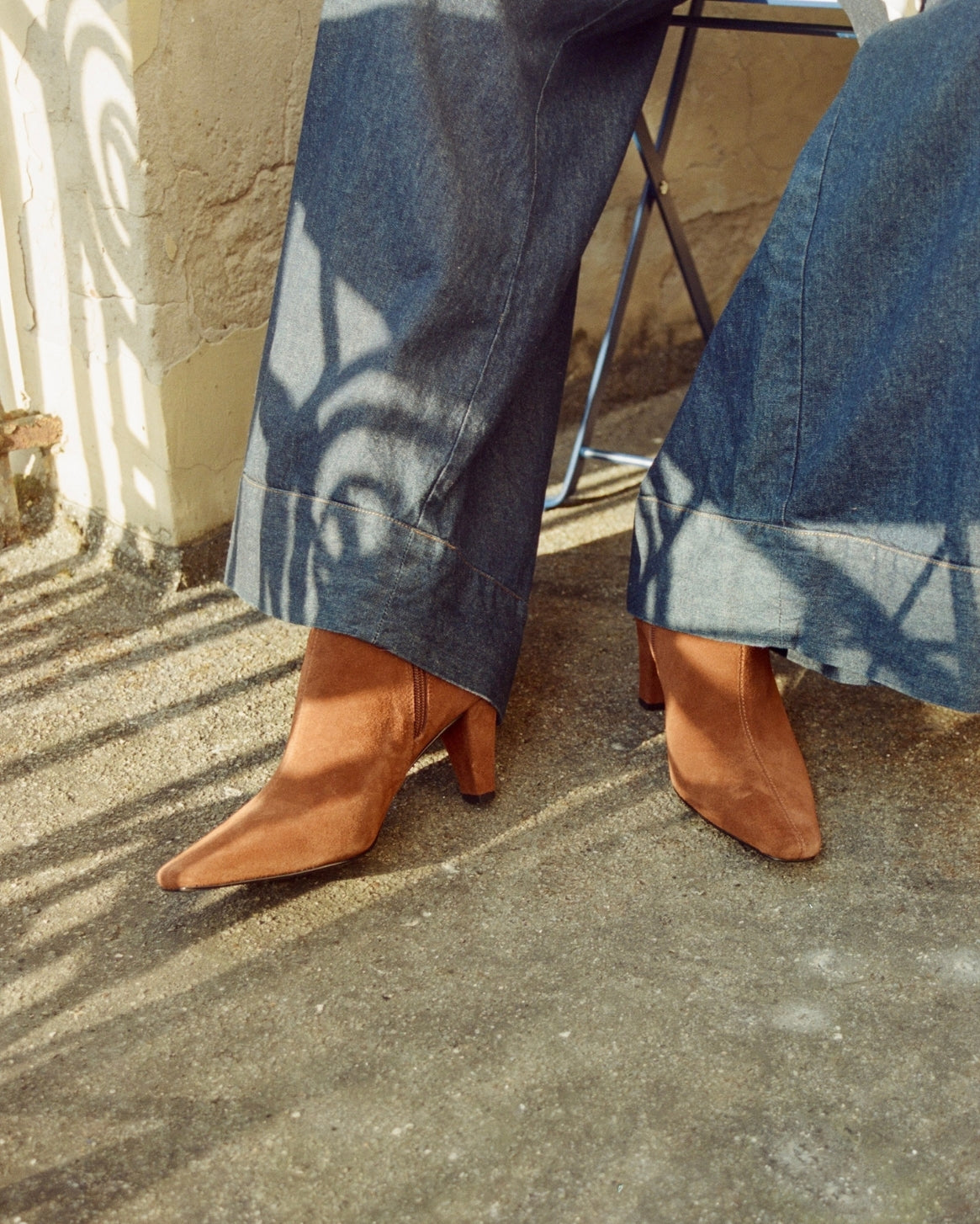 A person wearing blue wide-leg jeans and tan pointed-toe heeled ankle boots, seated outdoors on a concrete surface with dappled sunlight and shadows.