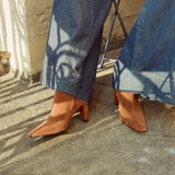 A person wearing blue wide-leg jeans and tan pointed-toe heeled ankle boots, seated outdoors on a concrete surface with dappled sunlight and shadows.