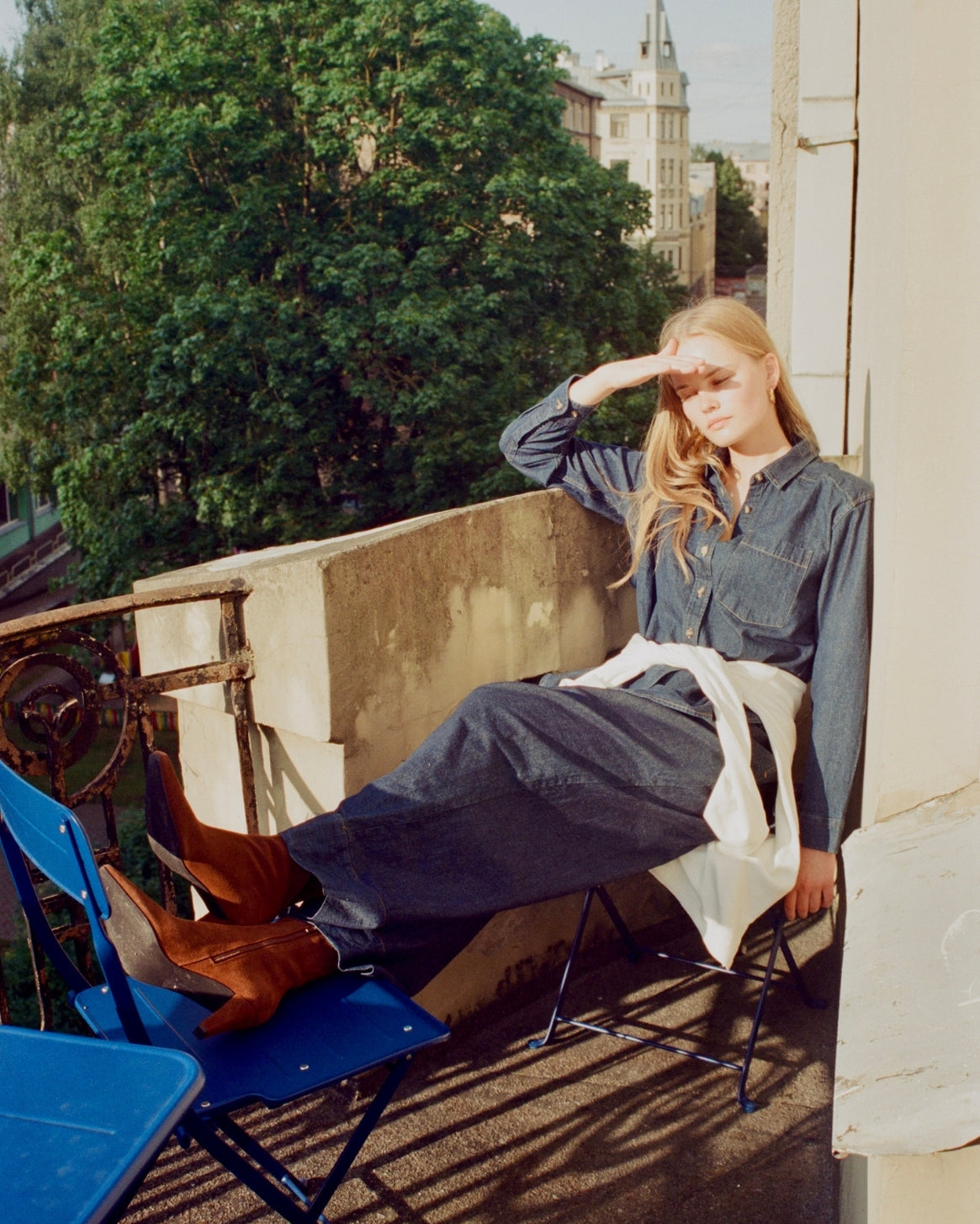 A woman sits on a folding chair on a balcony, wearing a dark denim shirt and wide-leg denim pants with a white shirt tied around her waist. She is wearing brown suede boots and shielding her eyes from the sun.