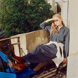 A woman sits on a folding chair on a balcony, wearing a dark denim shirt and wide-leg denim pants with a white shirt tied around her waist. She is wearing brown suede boots and shielding her eyes from the sun.