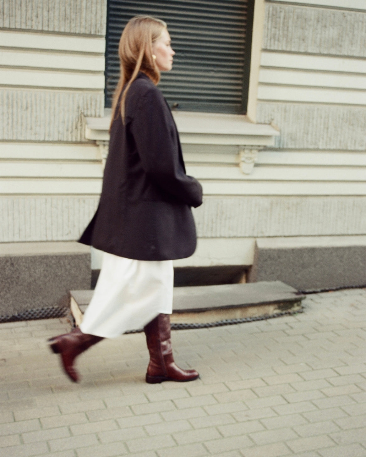 A woman walking in profile wearing a dark oversized blazer, a white midi skirt, and dark brown knee-high leather boots, set against a gray stone building background.
