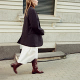 A woman walking in profile wearing a dark oversized blazer, a white midi skirt, and dark brown knee-high leather boots, set against a gray stone building background.