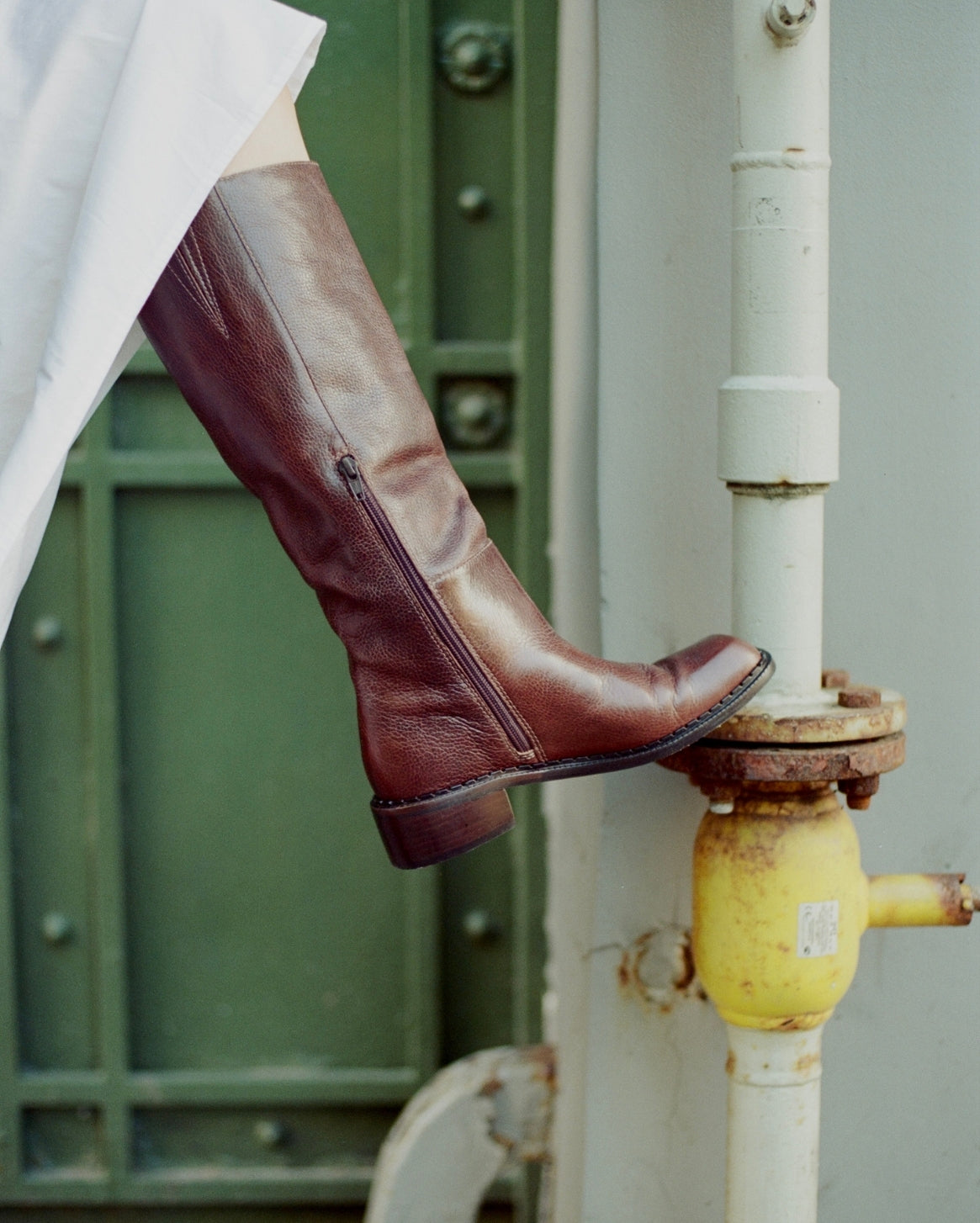 A person's leg wearing a dark brown leather knee-high boot with a side zipper, resting on a yellow fire hydrant next to a white pipe in front of a green door.