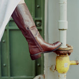 A person's leg wearing a dark brown leather knee-high boot with a side zipper, resting on a yellow fire hydrant next to a white pipe in front of a green door.