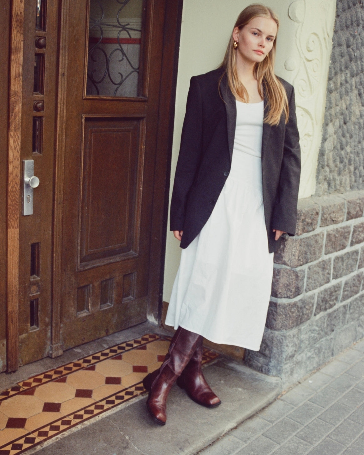 A woman stands on a doorstep wearing a white midi dress, a black blazer, and dark brown knee-high boots. She is posed in front of a rustic wooden door and stone wall.