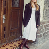 A woman stands on a doorstep wearing a white midi dress, a black blazer, and dark brown knee-high boots. She is posed in front of a rustic wooden door and stone wall.