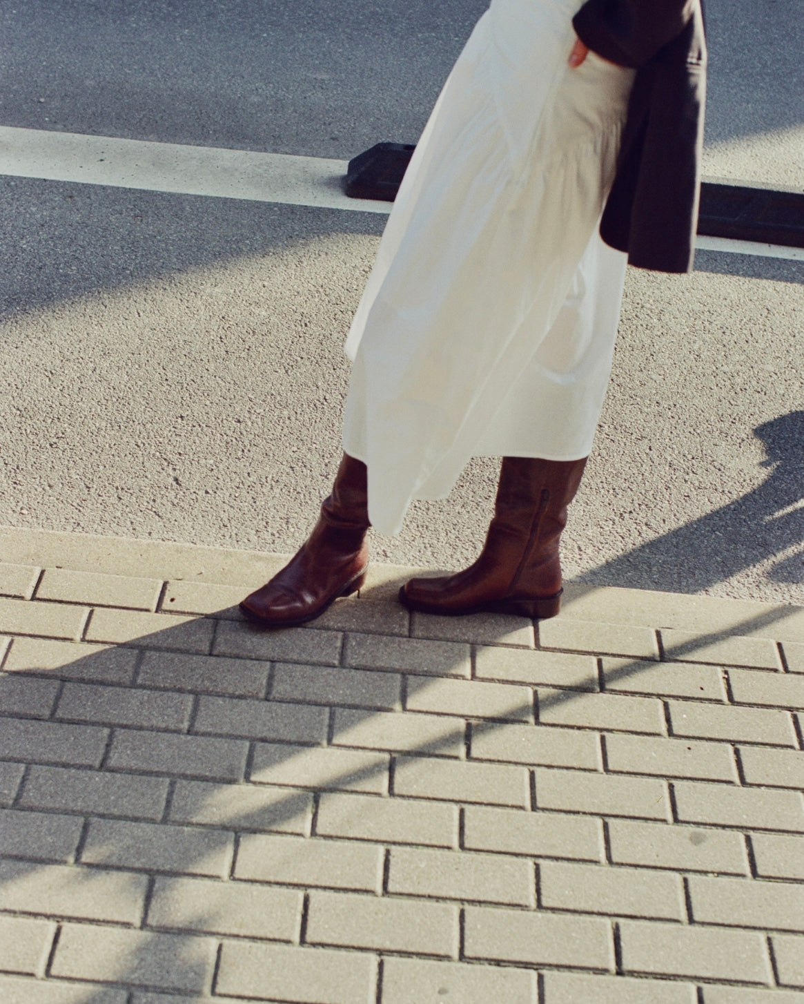 A person outdoors on a paved street corner wearing a long white skirt, dark brown knee-high boots, and carrying a dark shoulder bag.