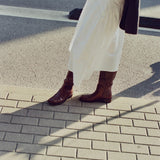 A person outdoors on a paved street corner wearing a long white skirt, dark brown knee-high boots, and carrying a dark shoulder bag.