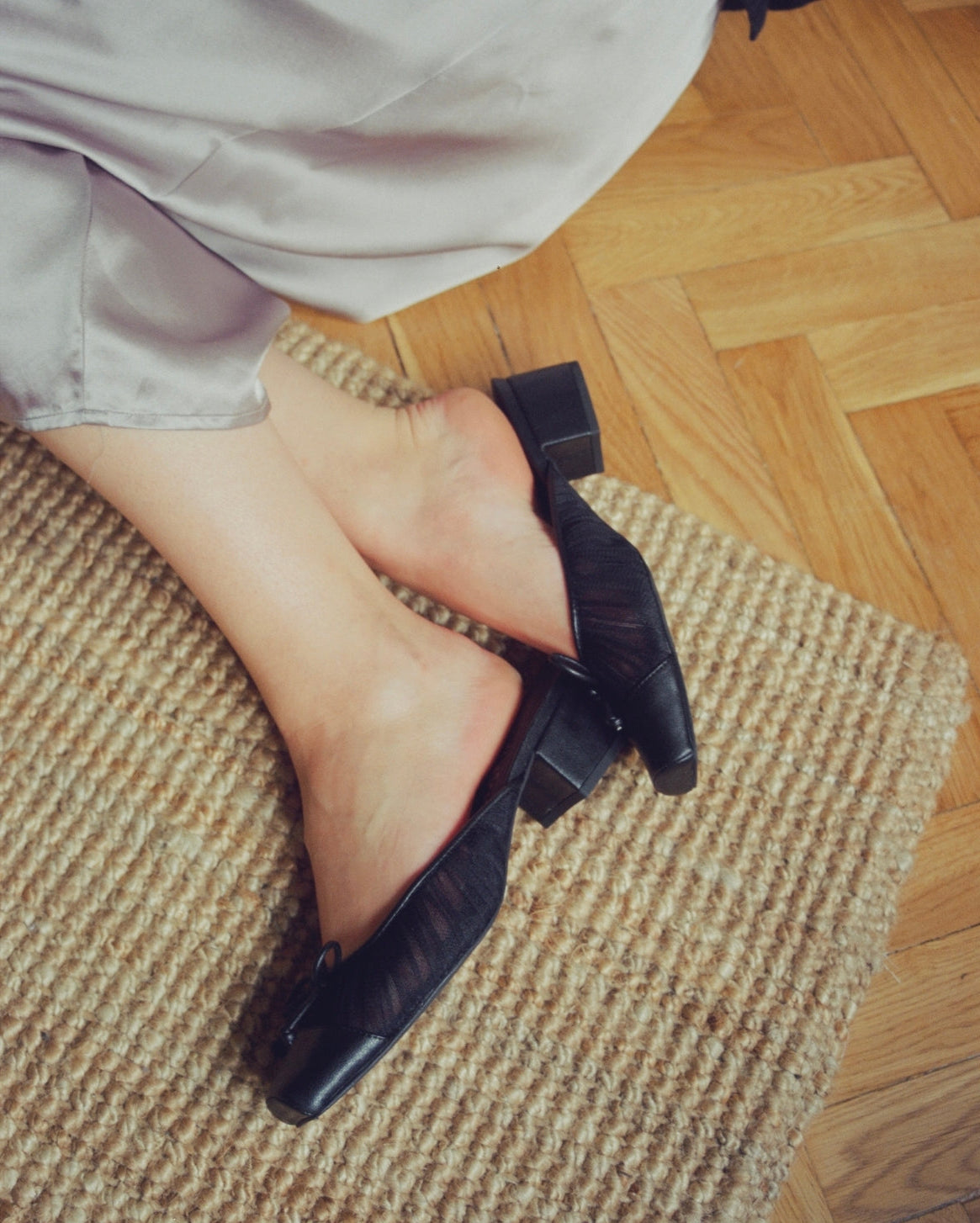 A person wearing black block-heel mules sits on a woven rug over a wooden floor, with the hem of a light-colored dress visible.