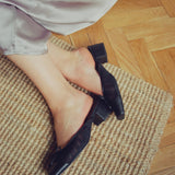 A person wearing black block-heel mules sits on a woven rug over a wooden floor, with the hem of a light-colored dress visible.