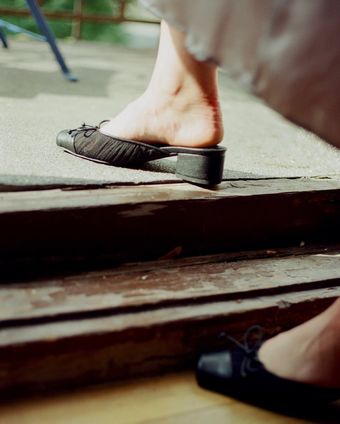 A close-up lifestyle shot of a person's bare foot stepping down rustic wooden steps into black low-heeled mules with bow details.