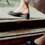 A close-up lifestyle shot of a person's bare foot stepping down rustic wooden steps into black low-heeled mules with bow details.