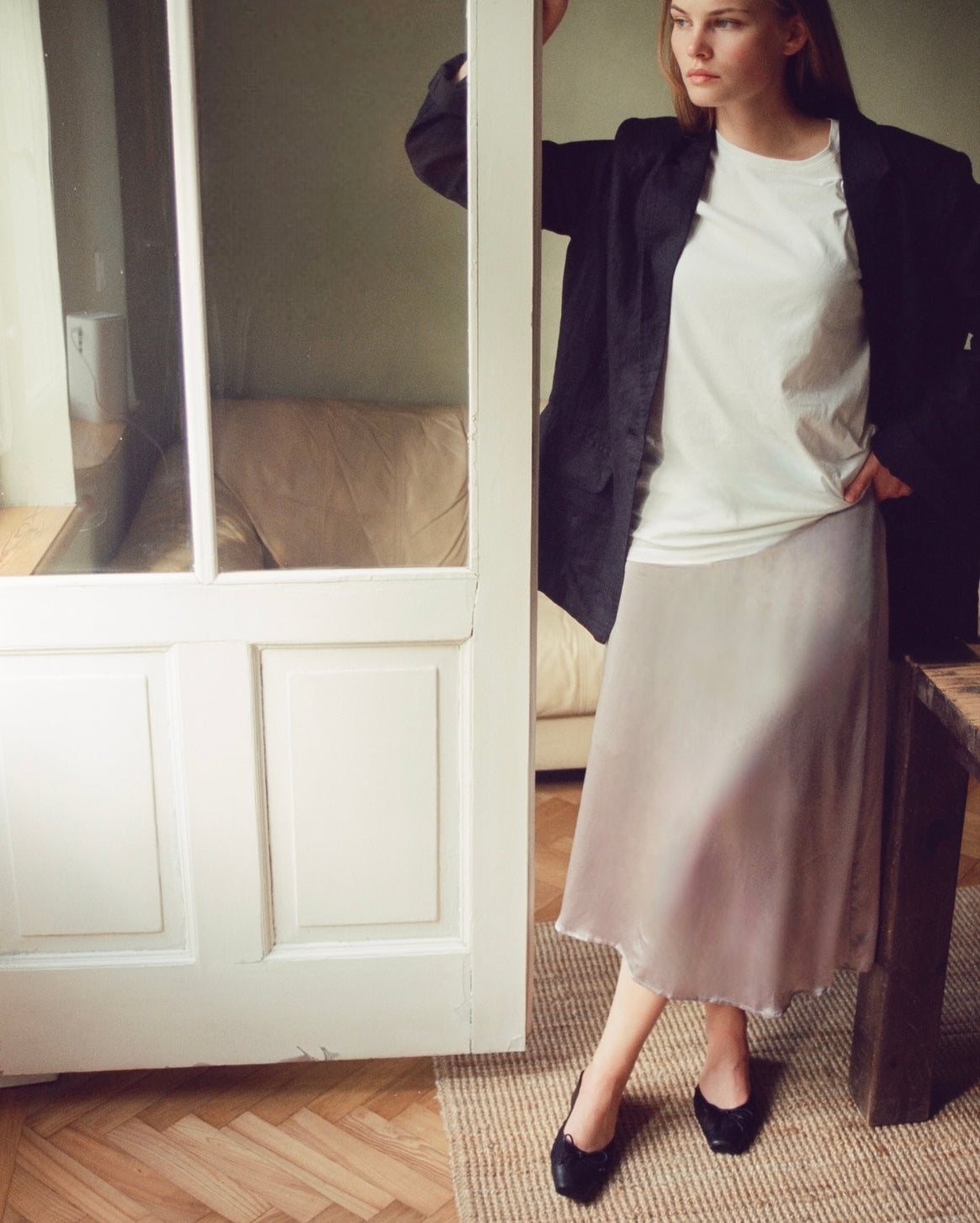 A woman models a light beige midi skirt with a raw hem, styled with a white t-shirt, a black blazer, and black heeled mules in a room with a wooden floor and a white paneled door.