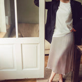 A woman models a light beige midi skirt with a raw hem, styled with a white t-shirt, a black blazer, and black heeled mules in a room with a wooden floor and a white paneled door.