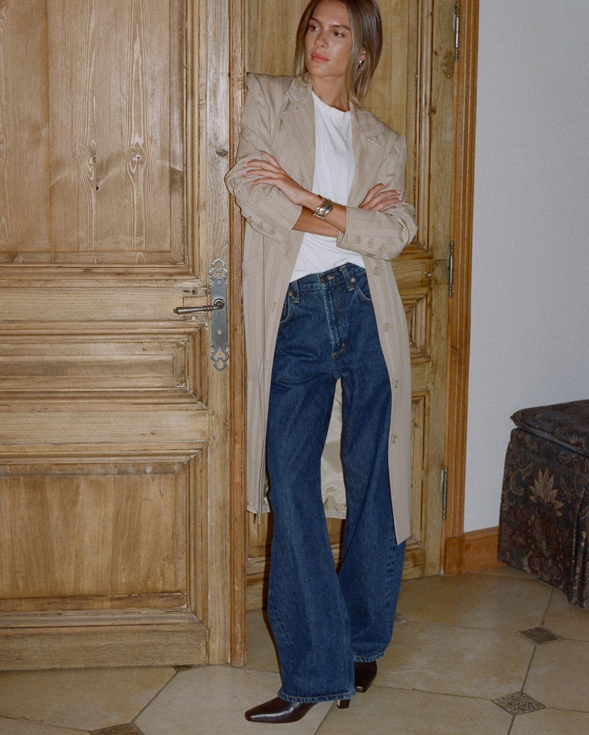 A woman stands in a doorway wearing a beige trench coat over a white top and dark blue wide-leg jeans, with brown shoes. The setting features a rustic wooden door and tiled floor.