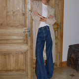 A woman stands in a doorway wearing a beige trench coat over a white top and dark blue wide-leg jeans, with brown shoes. The setting features a rustic wooden door and tiled floor.