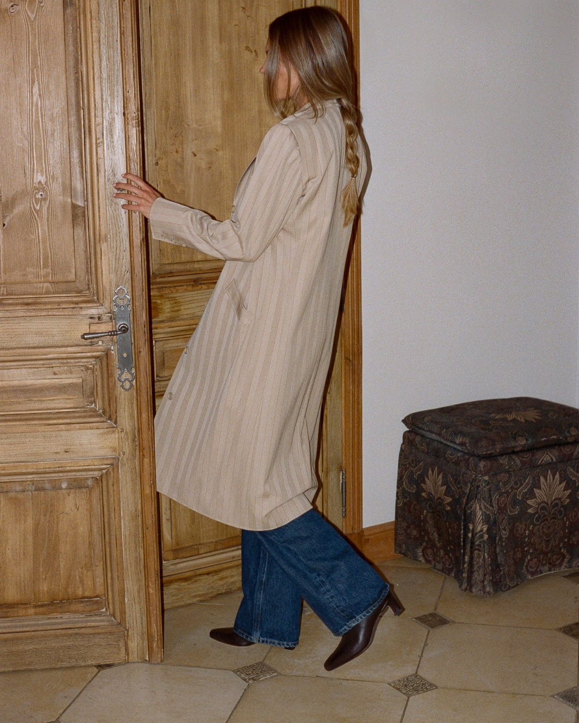 A woman wearing a long beige striped tunic shirt, blue jeans, and brown boots stands in a doorway next to a rustic wooden door, looking back over her shoulder.