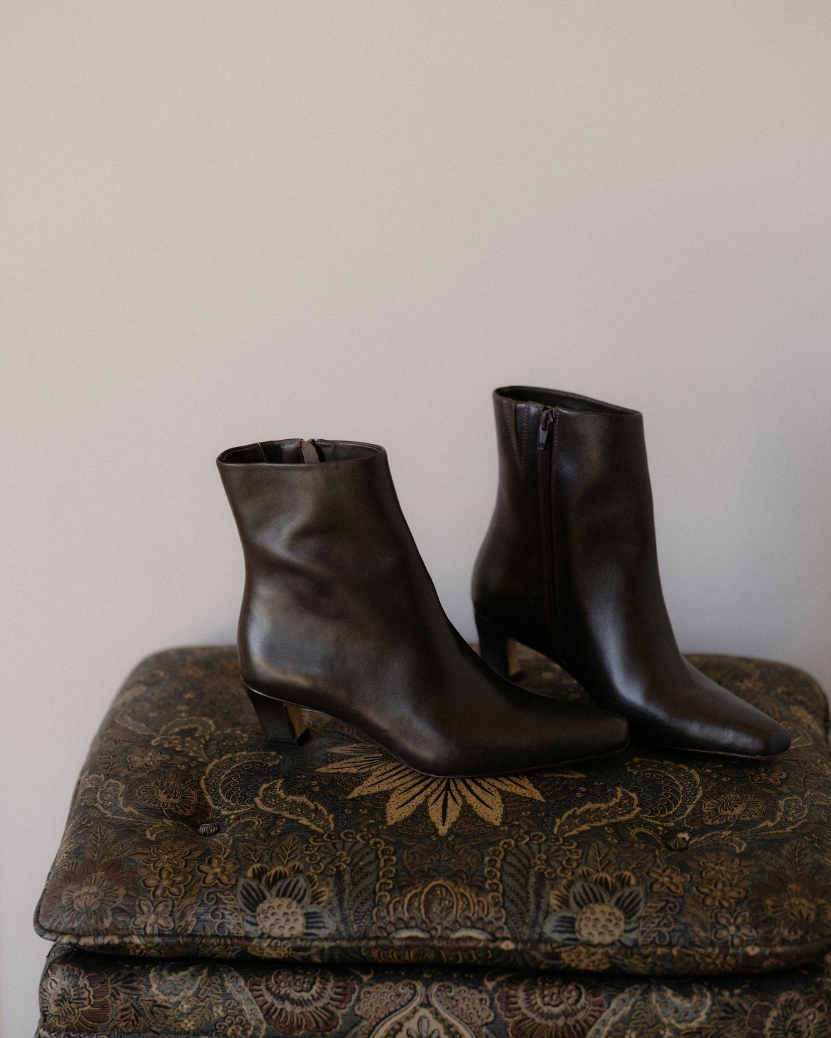 A pair of dark brown leather ankle boots with low kitten heels and side zippers, displayed on a patterned vintage ottoman against a plain beige wall.