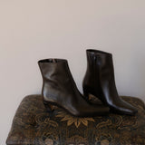 A pair of dark brown leather ankle boots with low kitten heels and side zippers, displayed on a patterned vintage ottoman against a plain beige wall.