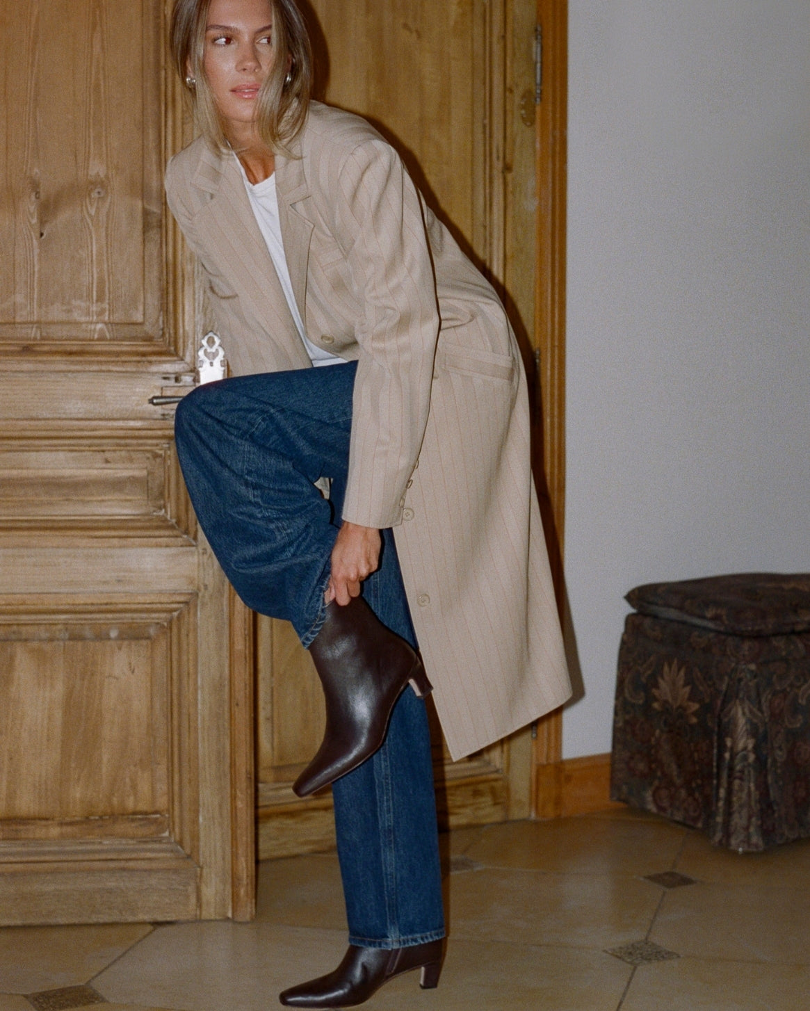 A woman in a beige trench coat, blue jeans, and a white top is shown from the waist down, lifting one leg to put on a dark brown leather ankle boot in an indoor setting with wooden doors.