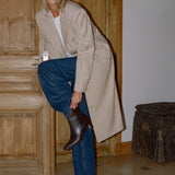 A woman in a beige trench coat, blue jeans, and a white top is shown from the waist down, lifting one leg to put on a dark brown leather ankle boot in an indoor setting with wooden doors.