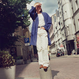 A person stands on a cylindrical bollard in the middle of a city street, wearing a loose white shirt, white wide-leg trousers, a blue open-front overshirt, and brown sneakers. Buildings, trees, and a cloudy sky are visible in the urban setting.