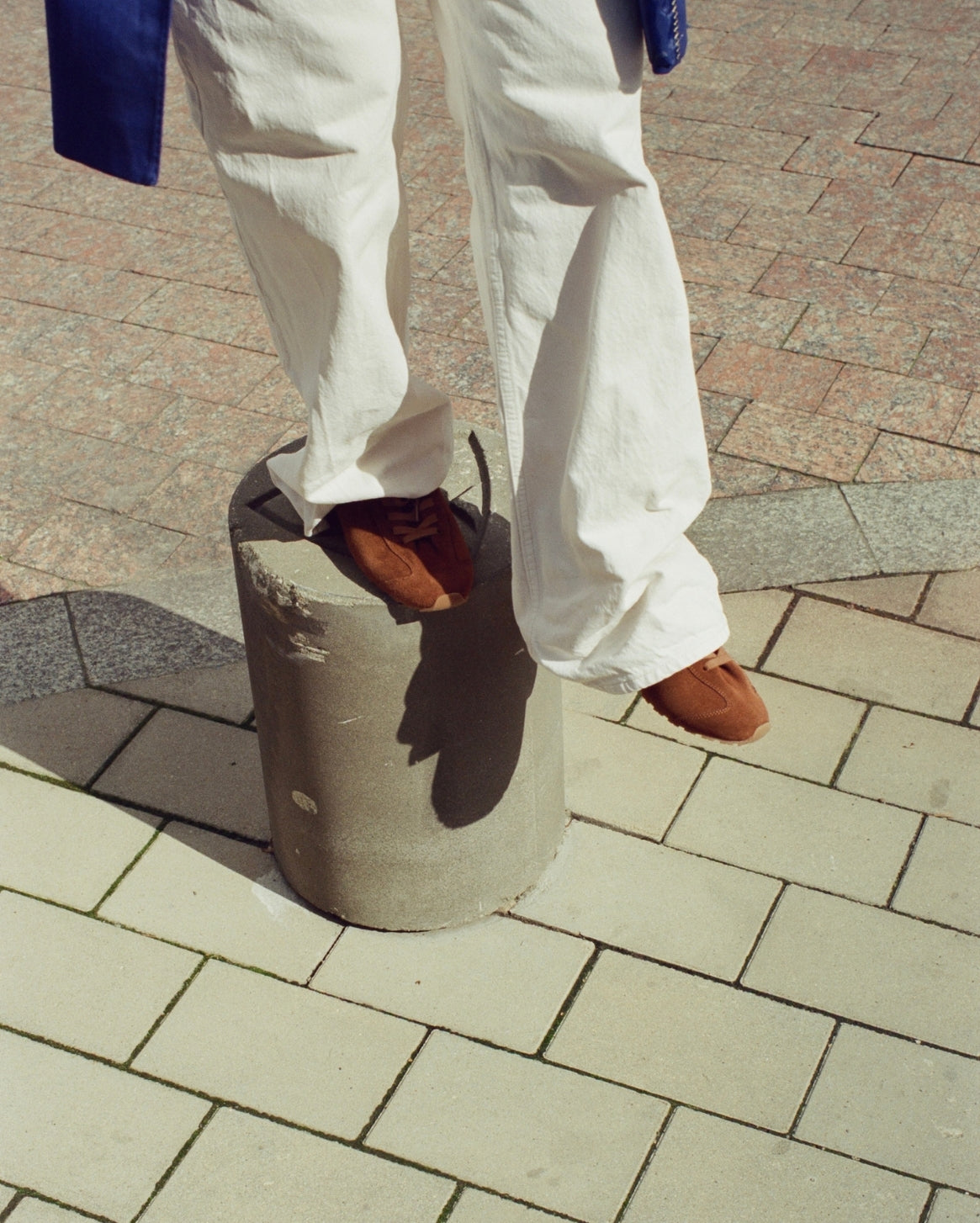 A person wearing off-white wide-leg trousers and brown suede sneakers stands on a cylindrical concrete block on an outdoor paved walkway.