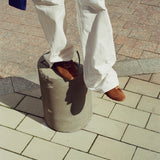 A person wearing off-white wide-leg trousers and brown suede sneakers stands on a cylindrical concrete block on an outdoor paved walkway.
