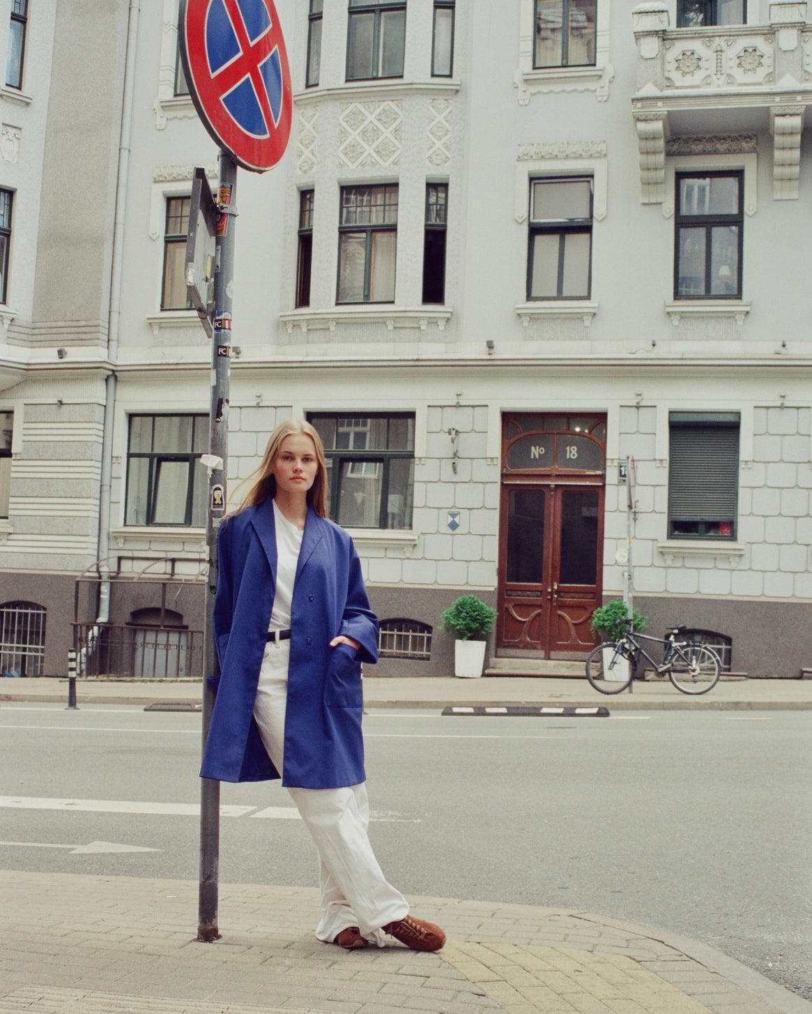 A woman stands on a city street corner wearing a bright blue tailored coat over a white top and white wide-leg trousers, with brown sneakers. A tall pole with a red-and-blue no-stopping sign is beside her, and a large light gray building with windows and a brown door is in the background.