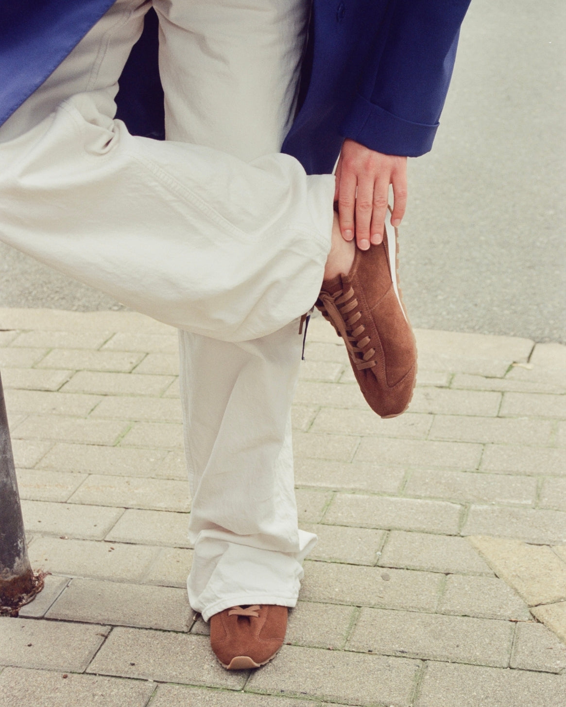 A person outdoors on a paved sidewalk wearing off-white wide-leg trousers, a navy blazer, and brown suede lace-up sneakers, with one leg raised and the shoe being held.