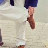A person outdoors on a paved sidewalk wearing off-white wide-leg trousers, a navy blazer, and brown suede lace-up sneakers, with one leg raised and the shoe being held.