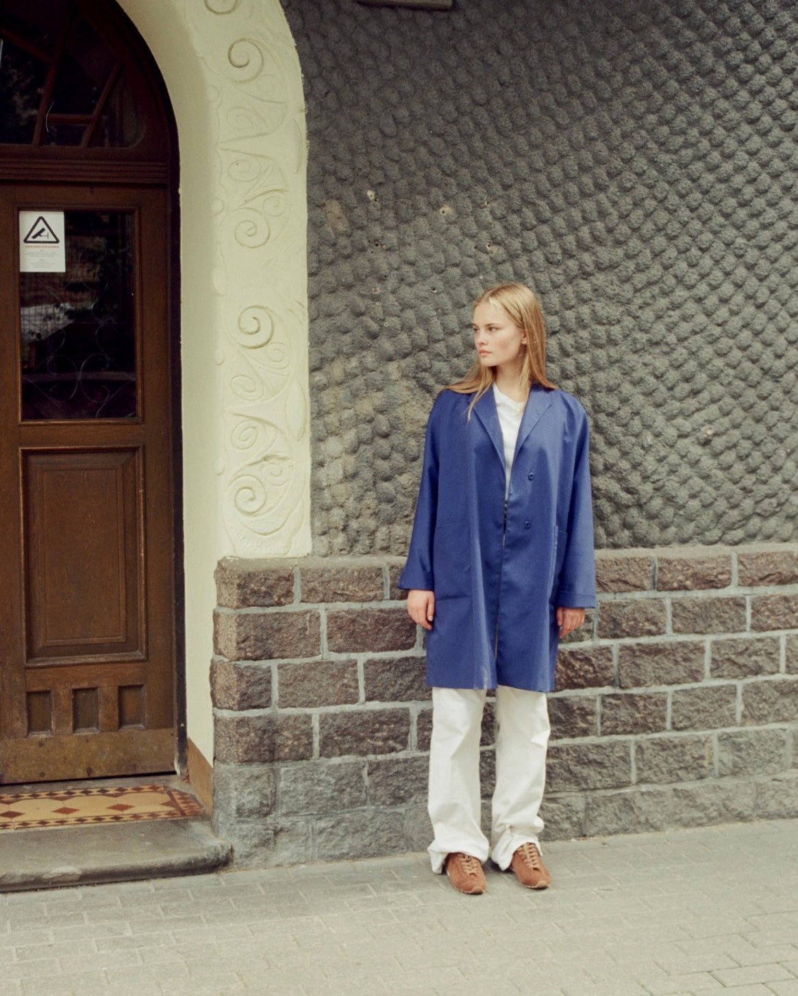 A woman stands outdoors against a textured stone wall and wooden door, wearing a long blue coat over a white top and white pants with brown sneakers.