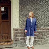 A woman stands outdoors against a textured stone wall and wooden door, wearing a long blue coat over a white top and white pants with brown sneakers.