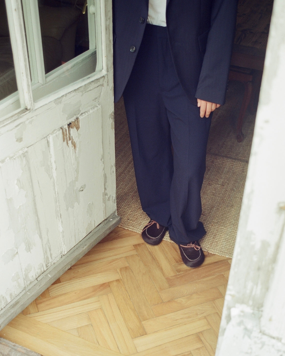 A person wearing a dark navy wide-leg suit with a white shirt and dark clog slippers, standing in a doorway with a light wood herringbone floor and a distressed white door frame.