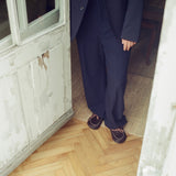 A person wearing a dark navy wide-leg suit with a white shirt and dark clog slippers, standing in a doorway with a light wood herringbone floor and a distressed white door frame.