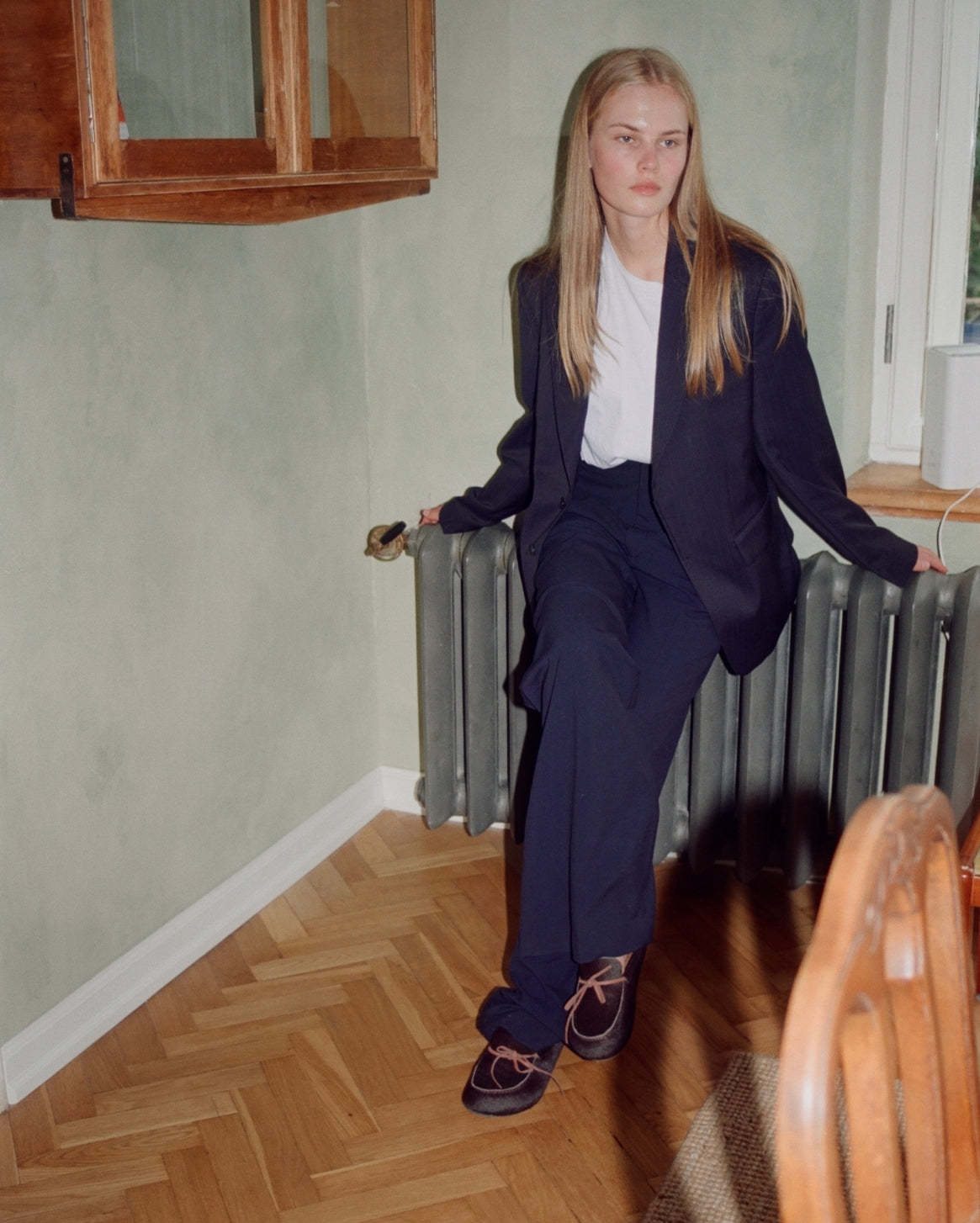 A woman with long blonde hair sits on a gray radiator in a room with pale green walls and a herringbone wood floor. She is wearing a dark navy blue tailored blazer and matching trousers over a white t-shirt, paired with dark brown lace-up clog slippers.