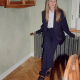 A woman with long blonde hair sits on a gray radiator in a room with pale green walls and a herringbone wood floor. She is wearing a dark navy blue tailored blazer and matching trousers over a white t-shirt, paired with dark brown lace-up clog slippers.
